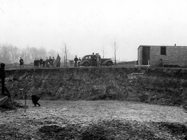 Excavations in Cross Flatts Park in November 1942. A workman stands in the large hole, where the front of a digger is visible. On the rim of the hole are members of the public and a parked car.