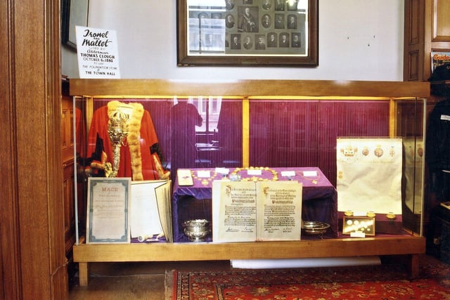View of the display cabinet, bought by Morley Council to display the civic regalia, in the small banqueting hall of Morley Town Hall pictured in October 1997. On the left is an aldermanic robe, the borough mace and a description of the mace. Next to this is the signature book for visitors to the Town Hall. Also seen is the Freedom-of-the-Borough silverware, the Mayor and Mayoress's chain, the deputy's chains, the granting of Morley's coat-of-arms and some coinage of the realm. The trowel and mallet mentioned on the notice at the top of the cabinet are to be found just in front of the visitors signature book.