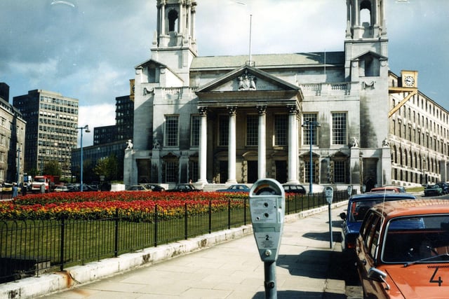 Leeds Civic Hall seen from Portland Crescent. Flower beds can be seen in Mandela Gardens in front. On the left is Calverley Street with Leeds General Infirmary and Leeds Polytechnic visible. Cars are parked beside parking meters in the foreground.