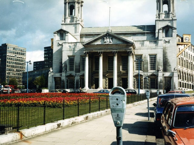 Leeds Civic Hall seen from Portland Crescent. Flower beds can be seen in Mandela Gardens in front. On the left is Calverley Street with Leeds General Infirmary and Leeds Polytechnic visible. Cars are parked beside parking meters in the foreground.