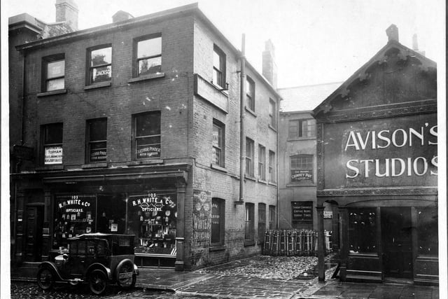 Albion Street in July 1927.  R.H. White, opticians. Upper floors occupied by various companies, logos on window for 'Brotherton Topham Estates and Insurance Agent, Rent and Debt collectors', 'Arthur Pinder, Motor Assessor'. On side wall, White has painted sign. In the yard at number 105 are The Patent Chimney Company with window notice and Allied News Ltd list in lower window Sunday Chronicle, Empire News and Sunday Times. 105 was also used by the Antique Reproduction Silver plate manufacturers. Number 107, studio of Edwin Avison photographer, with decorative wooden trim.