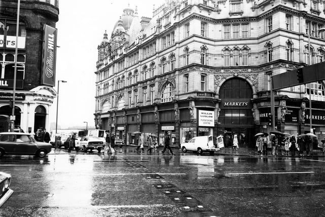 Looking east from Vicar Lane towards Ludgate Hill, with Kirkgate Market on the right. On the Ludgate Hill side of the market entrance is Willis Ludlow, department store, and on the Vicar Lane side is Barker's children's wear and baby linen. The National Westminster Bank can be seen on the right of the photograph. The photo was taken on a wet day in January 1979 and there are many people with umbrellas.