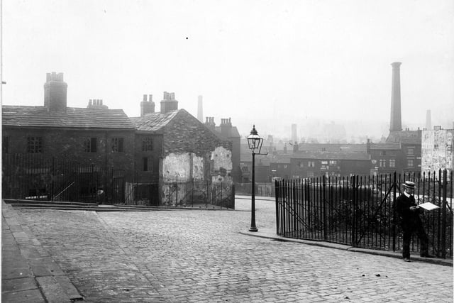 Taken during the Quarry Hill improvements in August 1906. Showing on the left a row of derelict terraced houses and a railed garden area on the right. There is a man, possibly a surveyor on the right. In the background are the shops and houses of Mabgate. There is an advertising hoarding on the left, full of unidentified posters. Beyond that is an unclear view of the roofs and chimneys of Leeds.