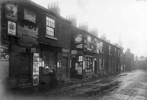 A parade of shops on Domestic Street pictured in November 1929. On the right is 28 Domestic Street - newsagents owned by Mrs Ethel Hartley. Next is Cross Atkinson Street, then 24 Domestic Street - a small shop owned by William Lindsay. Advertisements are plastered over the fronts of both shops. St. Matthew's Church is visible in the distance.