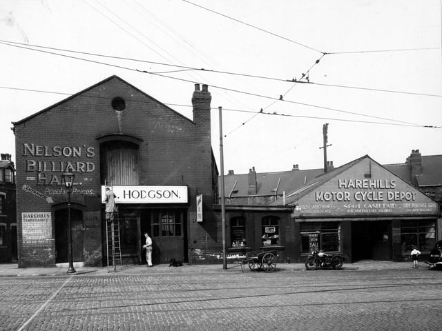 The west side of Harehills Road showing Nelson's Billiard Hall, Hodgson's and Harehills Motorcycle Depot with a young boy looking in the window and a motorcycle outside. Man on ladder painting sign. Wooden cart, dog and tramlines visible on road. Pictured in August 1947.