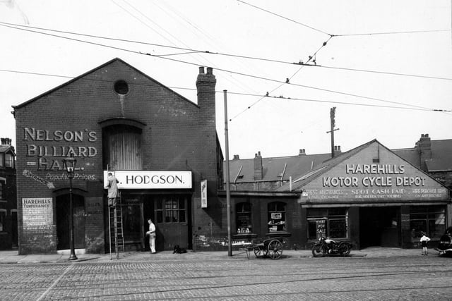 The west side of Harehills Road showing Nelson's Billiard Hall, Hodgson's and Harehills Motorcycle Depot with a young boy looking in the window and a motorcycle outside. Man on ladder painting sign. Wooden cart, dog and tramlines visible on road. Pictured in August 1947.