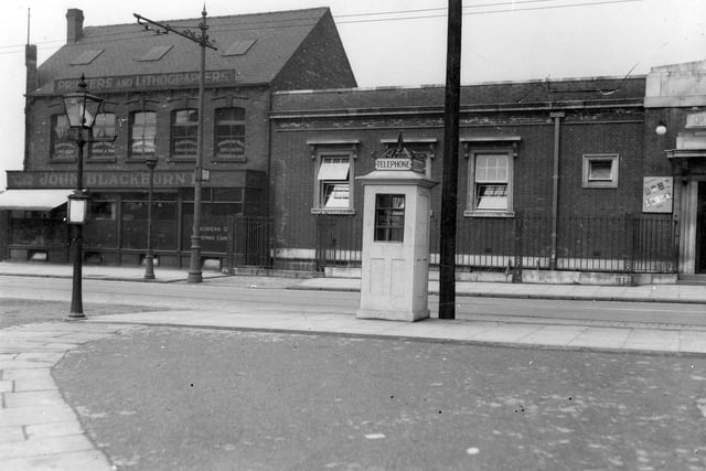 Telephone box on Cardigan Road at the junction of Thornville Road. Behind the telephone is Burley Library. Adjoining Burley Library is John Blackburn Ltd, printers and lithographers. On the left hand side of the photo is a street lamp. Pictured in June 1936.