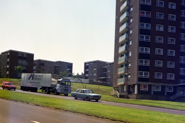 A photograph of a High Street Transport lorry driving down Oak Tree Drive in Gipton. This is locally known as ‘Fairway Hill’, so named after the public house that once stood at the top of the hill. There is a tower block on the left side of the road and several blocks of low-rise council housing. There are also two cars driving down the road. This is a Burton's company photograph. Pictured in June 1973.