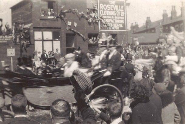 A carriage with the King and Queen in procession along a main road (possibly Roseville Road) in August 1933. A street procession and crowds waving. A background building with wall advert or sign depicts The Roseville Clothing Co. Ltd.