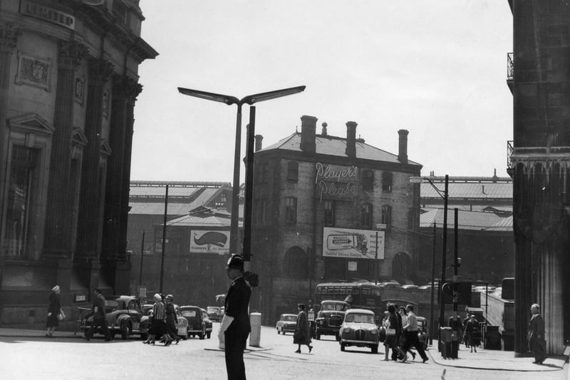 A police officer directs the traffic on Boar Lane at the busy junctions with Bishopgate Street where cars are visible towards the left, and Wellington Street, far right. On the of Boar Lane and Bishopgate Street the rounded decorative building seen far left is the former Yorkshire Banking Company, the Midland Bank Ltd. Leeds City Station can be seen between the buildings in the background, and on the far right is part of the Queens Hotel.