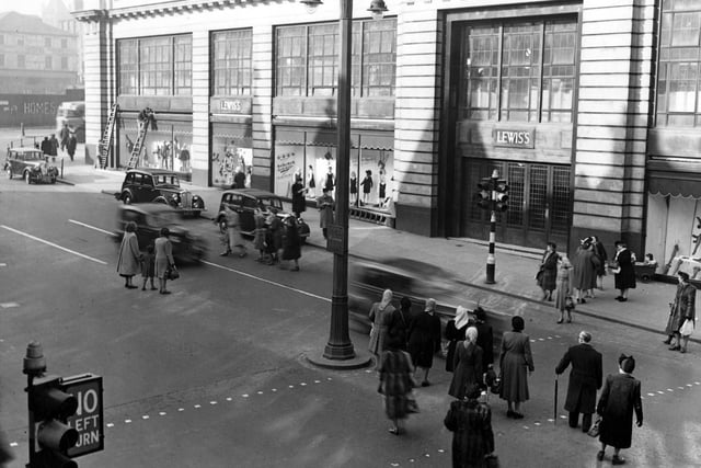March 1949 with Lewis's (Leeds) Ltd. department store on the north east side of The Headrow. Cars are parked outside. Shoppers are crossing the road. On the left, two men are working up ladders. Some graffiti can be seen on Wade Lane. A double lampost and traffic lights are prominent.