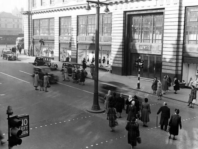 March 1949 with Lewis's (Leeds) Ltd. department store on the north east side of The Headrow. Cars are parked outside. Shoppers are crossing the road. On the left, two men are working up ladders. Some graffiti can be seen on Wade Lane. A double lampost and traffic lights are prominent.
