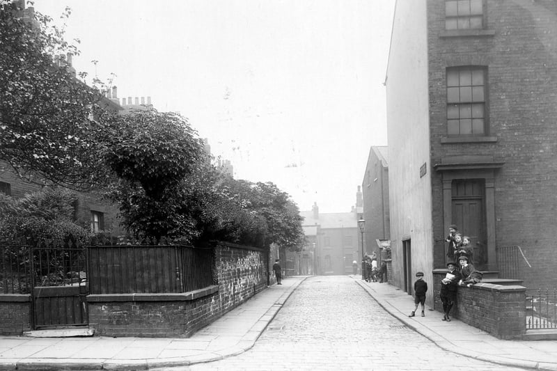 South Carnaby Street, looking south-east towards Cankerwell Lane in August 1913. Intersection of streets, houses of various sizes and styles. Left side, garden walls and fences with trees and shrubs. On right, groups of children posing on wall, dressed in period style.