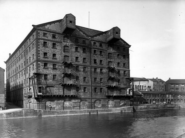 A view of British Waterways warehouse seen from River Aire in September 1950. The building is a large eight storey building with an enclosed hoist mechanism on the top floor. Each floor has a door and a balcony from which goods could be raised or lowered. There are more warehouse buildings to the right along the riverside.