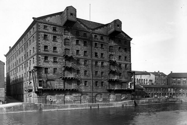 A view of British Waterways warehouse seen from River Aire in September 1950. The building is a large eight storey building with an enclosed hoist mechanism on the top floor. Each floor has a door and a balcony from which goods could be raised or lowered. There are more warehouse buildings to the right along the riverside.