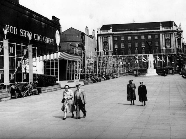 Coronation decorations on Victoria Gardens outside the Central Library and Art Gallery on The Headrow. The Art Gallery has 'God Save the Queen' on the wall. Along the length of the garden, site scaffolding has been put up, which is supporting banners and crests. A platform has been built in the centre. Looking towards Cookridge Street the war memorial can be seen, with flags of St. George.