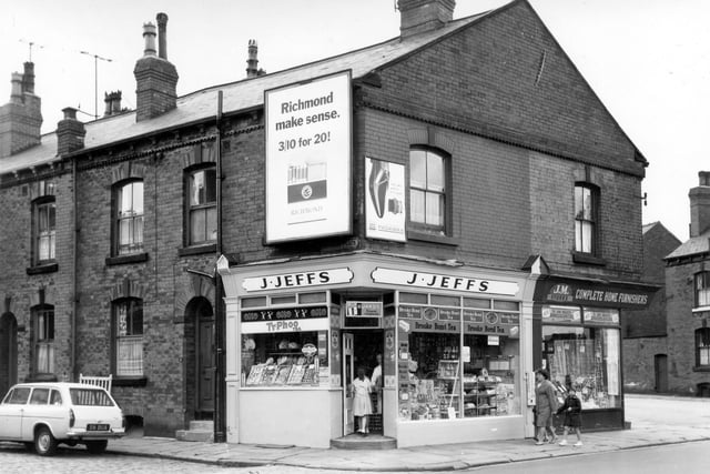 On the left is the back-to-back terraced houses of Jesmond Place. The shop in the gable end is number 103 Hunslet Hall Road a J. Jeffs grocery. The grocer stands in the doorway talking to two young girls while a mother walks another girl towards the shop. On the right is J.M. Stores, complete home furnishers selling, Axminsters and Wiltons carpets. On the far right is Jesmond Terrace.