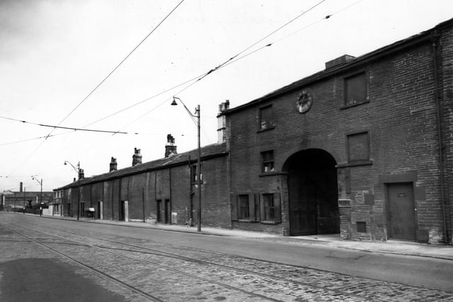 The entrance to Burley Mills on Kirkstall Road showing a large arched gateway. There is a clock above this. The ground floor windows have shutters. Along the road are the premises of Raleigh Industries Ltd., cycle makers. Pictured in September 1950.