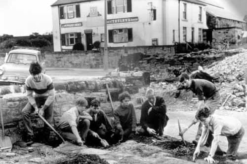 The Cardigan Arms provides a backdrop this this photo circa 1965.  Pupils from Woodkirk School are working with shovels and pick-axes under the supervision of their teacher. They are excavating an area in front of Woodkirk Church (St. Mary's) tower.