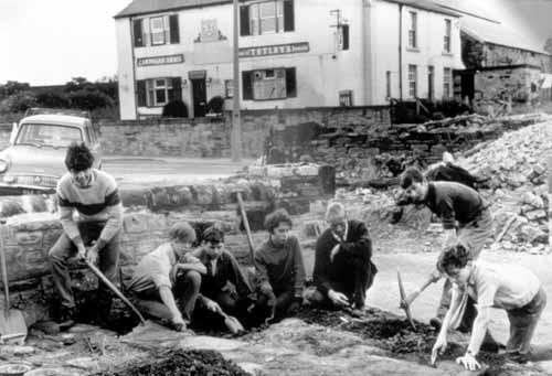 The Cardigan Arms provides a backdrop this this photo circa 1965.  Pupils from Woodkirk School are working with shovels and pick-axes under the supervision of their teacher. They are excavating an area in front of Woodkirk Church (St. Mary's) tower.