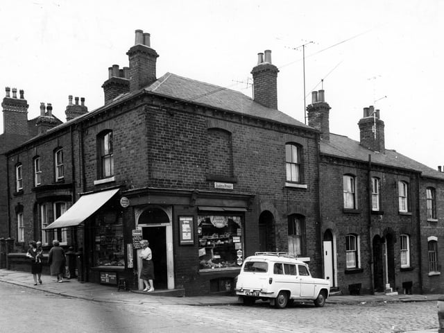 A shop keeper stands at the door to greet customers on the corner of Malvern Road to the junction with Algeria Street. The shop sells groceries and 'local grown caulis, cabbages, carrots and peas.'