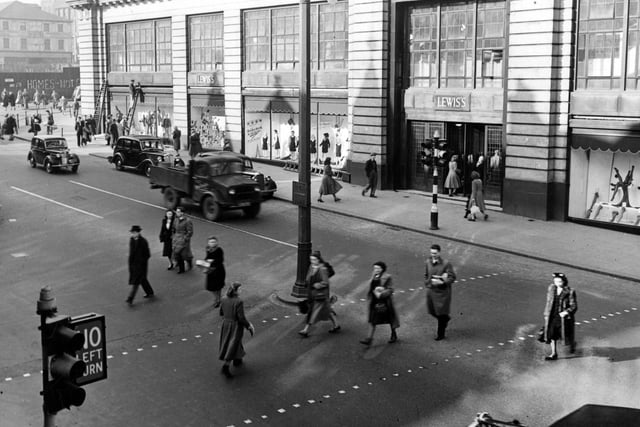 A view of Lewis's (Leeds) Ltd. department store on The Headrow. Cars and a lorry are on the road which pedestrians are crossing. Some graffiti can be seen on Wade Lane on the left. A man works up a ladder. A lampost and traffic lights are visible. Pictured in March 1949.