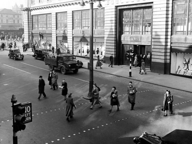 A view of Lewis's (Leeds) Ltd. department store on The Headrow. Cars and a lorry are on the road which pedestrians are crossing. Some graffiti can be seen on Wade Lane on the left. A man works up a ladder. A lampost and traffic lights are visible. Pictured in March 1949.