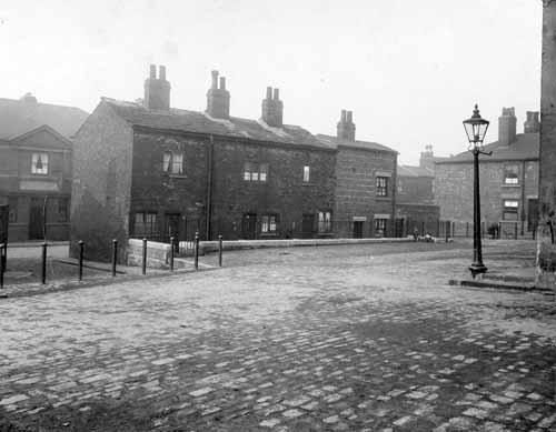 The square on Dolphin Street pictured in March 1930. Terrace houses to left are on Richmond Road, a group of children and a dog play at the back of the square. Gas lamp post to the right, houses beyond it are on Dolphin Street. Metal fences in front of the wellhouses in the foreground of the picture.