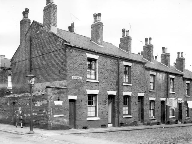 The first door on the left gives access to a one storey building in poor condition which houses toilet facilities for these back-to-backs on Algeria Street. A gas street lamp stands on the corner of the street and a lady walks along Kiln Street.