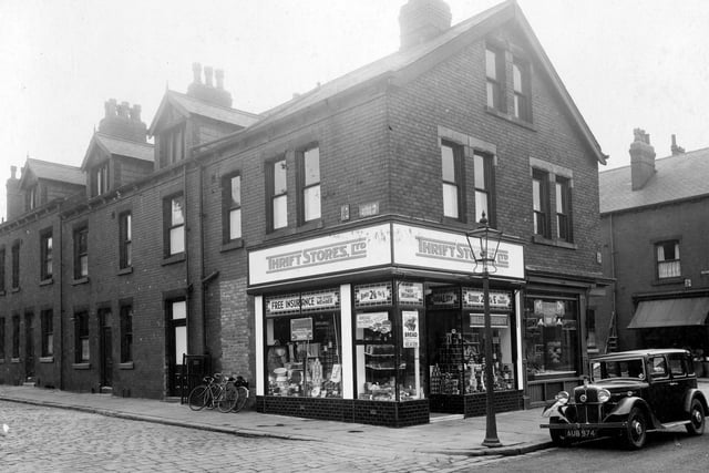 The Thrift store on Morris lane at the junction with Station Parade in August 1935. The chain of shops was founded by James Popplewell of Holbeck. There are displays of hardware, tinned goods, bread and cakes in the windows. The exterior of the shop has been upgraded with new signs and tiling under the windows. To the right is William Greaves butcher. This street is Glebe Avenue. There are bicycles outside the Thrift Store and a car in the road.