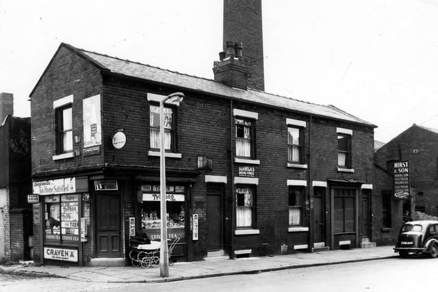 A corner grocers on Jack Lane in August 1963. Bubble gum machines flank the door and windows while a coach built pram waits outside the door. On the right are residences with the Hirst and Son (Leeds) Ltd, electrical engineers, electrical contractors advertising Motor rewinds on the far right. A man stands in the doorway while a van, reg: CDN 449 is parked on the street. In the background behind the building is a chimney belonging to the vehicle builders.