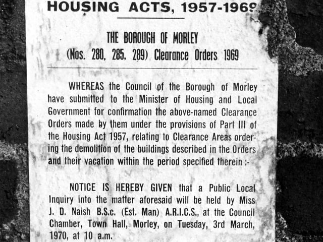 A notice on the wall of a house on Parker Street in Morley. The notice is headed 'Housing Acts, 1957-1969' and relates to the Clearance Orders' of 1969. It advertises a public inquiry into the demolition of the buildings to be held at the Council Chamber, Town Hall, Morley, tuesday 3rd March, 1970 at 10am.