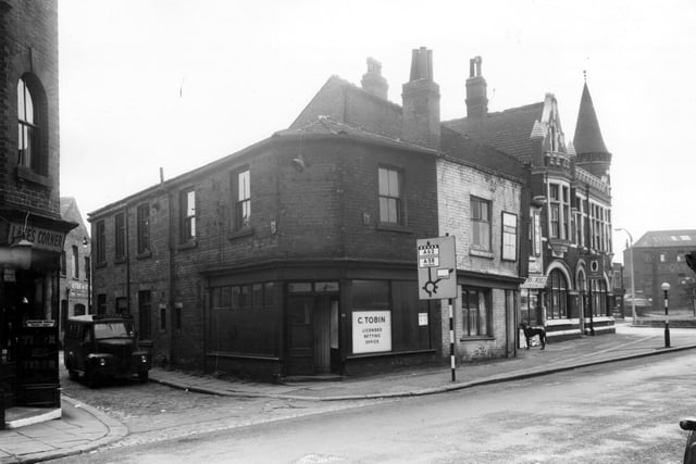 Holbeck Lane in March 1965.  On the left edge of this view, is the doorway of Lake's corner, a grocers run by Frederick Lake. The entrance to Stead Street follows to the right where a van for John Cooke and Son of Huddersfield is parked. This firm were concrete engineers and had works on Coleman Street nearby. Continuing right is C. Tobin's licensed betting office. Outside this shop is a road sign directing motorists along Wortley Lane to Huddersfield and Halifax and along Spence Lane to Armley. Towards the right the crenellated turret of the Volunteer public house is visible.