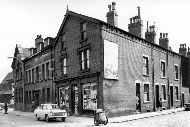 On the far left just visible is St Peter's Church consecrated July 1868, then Dewsbury Road with the police station, which opened in July 1903. Next on the right of the police station is a childrens clothes shop taking up the gable end of Jesmond Place. An advertising hoarding on the side of the shop promotes New Zealand Cheddar. A car, reg: 708 JOC is parked outside the clothes shop and a dust cart waits at the corner while its operator works on the far left as people walk along Hunslet Hall Road. A woman stands in the doorway of number 1 Jesmond Place on the right while a young boy stands in front of number 3. On the far right is a yard originally built to house the shared outside toilet.