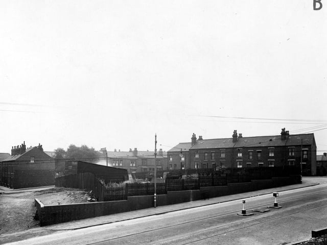 Belle Isle Road in March 1946. with tram lines and a traffic island. In the middle is a small piece of ground with some road mending equipment and rubbish on it. There is a wooden shed with a sloping roof, all surrounded by a wooden fence. A low, graduated brick wall runs along the main road. In the background are three rows of brick terraced houses. Electric cables run across the view, one is carrying a suspended light.