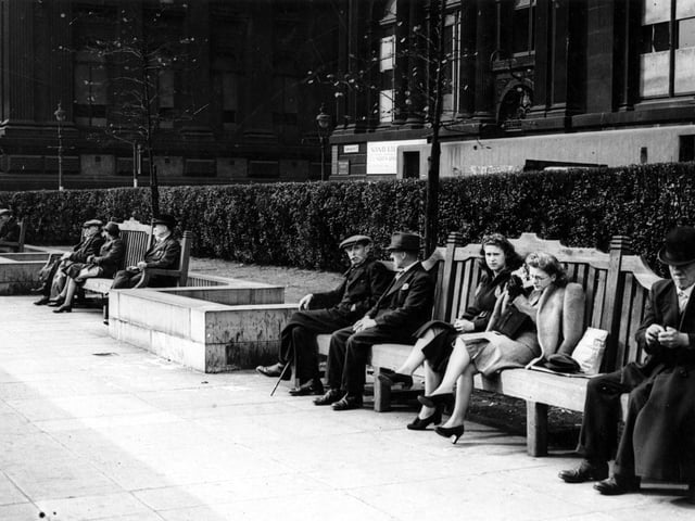 The Garden of Rest between The Headrow and Centenary Street looking west across Calverley Street to the Town Hall. In the foreground are long benches where people are sitting. Behind is a hedge, and further back, the Municipal Buildings (including the library). Air raid protection around ground floor of Municipal Building. Pictured in September 1943.
