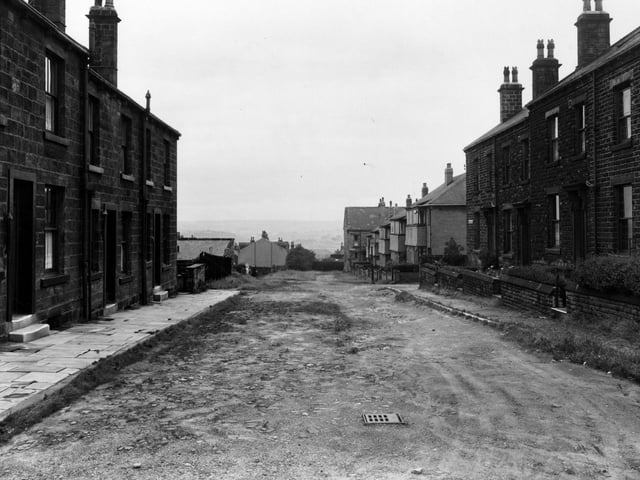 A view looking south down the Melbourne Grove. in August 1954. Houses at nos. 3-28 are visible. The road is unsurfaced but flat. A scenic view is visible in the background. Two people can be seen in the distance.