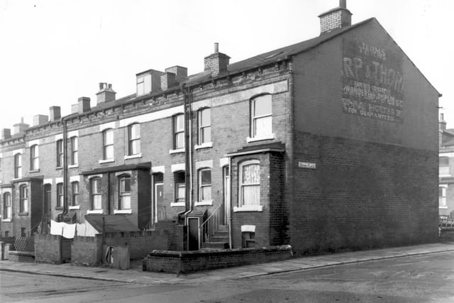 The gable end of back-to-back terraced houses on Ascot Street. Stairs lead up to first floor entrances and front yards containing washing lines are surrounded by short walls. On the wall of the Devon Place facing gable are the remains of painted advertisements. Pictured in October 1966.
