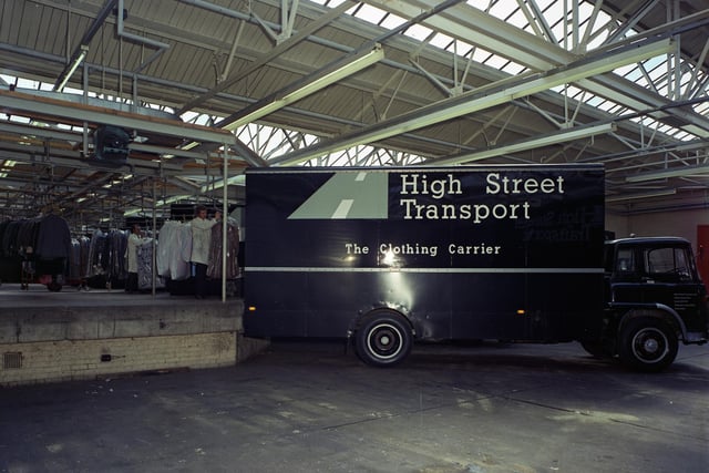A photograph of men loading garments on coat hangers into the back of a High Street Transport lorry in June 19i73. There are rows of clothes hung up on rails waiting to be loaded onto the lorry. The sign on the side of the lorry reads 'High Street Transport: The Clothing Carrier'. The photograph was possibly taken in Burton's clothing factory on Hudson Road.