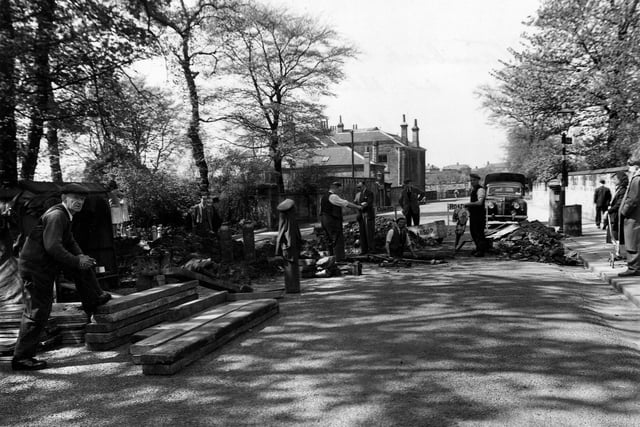 Road subsidence on Stainbeck Lane with maintenance work in progress. A car, a bus stop, workmen and onlookers are visible. Pictured in May 1953.