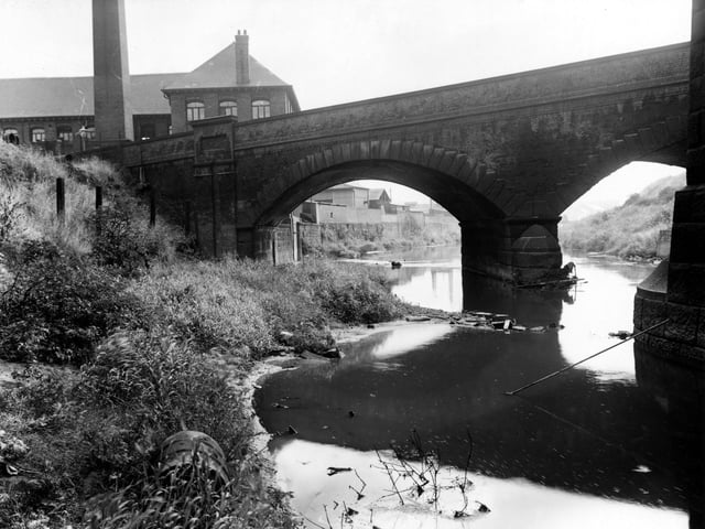 The bridge of Viaduct Road as it crosses the River Aire, looking east, in September 1955. The stone structure on the far right is one of the supports of the L.N.E.R. viaduct. Behind the bridge, to the left, is the factory and chimney of Charles F. Thackray Ltd., surgical instruments. The river bank is overgrown, with an old oil drum in the foreground.