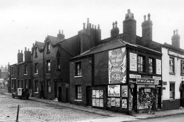To the left, at the top of Delph Street are the remains of White Rose Yard. Numbering on Delph Street begins here with 12 and moves right in descending order to 2, which is next to all the wall posters. This is the corner with Woodhouse Street. Number 204 is a newsagents and hardwaare shop, business of Mrs Getrude Moss. The shop window is full of goods and there are many advertisements for products and publications. To the right is 202. This is the White Rose public house selling Melbourne Brewery beers. Pictured in September 1959.