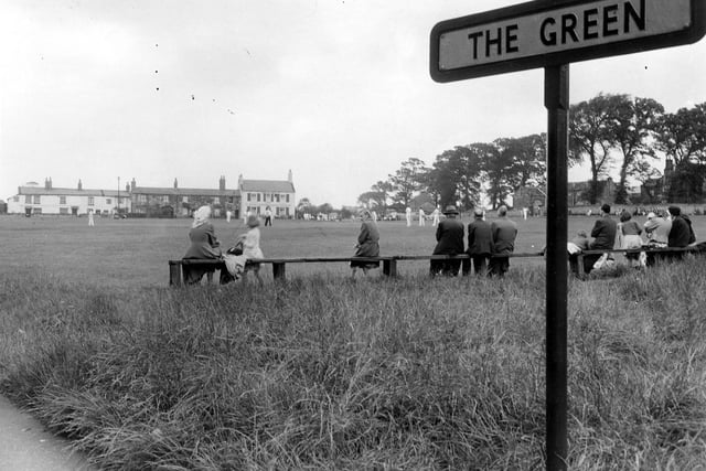 A cricket match in progress on The Green at Seacroft in 1961 supported by spectators seated on wooden benches. The view is from York Road across the Green to properties including the Cricketers' Arms public house (a large white-rendered building, left of centre). On the right, the old Seacroft Grange County Primary School is visible.