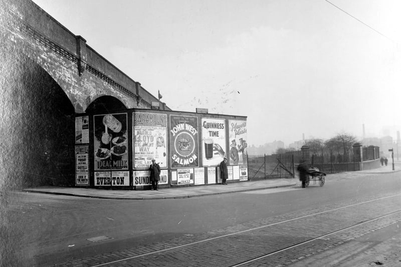 Kirkgate, where Cross York Street passes under the LNER viaduct, in April 1936. Two men in cloth caps and over coats are standing in front of free standing advertising hoarding.