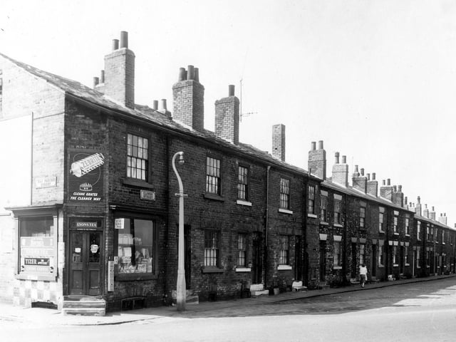 On the left of the image and in the foreground is New Princess Street with number 1 visible at the corner. This is a grocery advertising products including Zebrite polish, Tizer, Lyons coffee and tea and Senior Service cigarettes. On the right of the image is a row of back-to-back terraced houses numbers 1 to 19 Clowes Street.