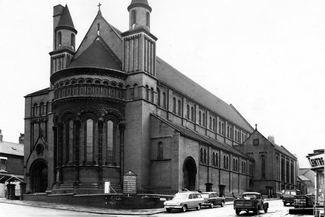 St Aidan's Church on Roundhay Road in April 1965 with Elford Place West on the right. Built in 1891-4 by R.J. Johnson & A.C. Hicks, St Aidan's joins Basilican form with Romanesque style. The image shows a double-apsed aisled basilica with a basilican chapel attached on the south eastern side. The interior of the church is famous for the large display of mosaics in the apse and on the concelli walls by Frank Brangwyn. Red brick and mosaic were chosen as construction materials as they were easy to maintain in Leeds polluted atmosphere. A large board in front of the church lists mass times.