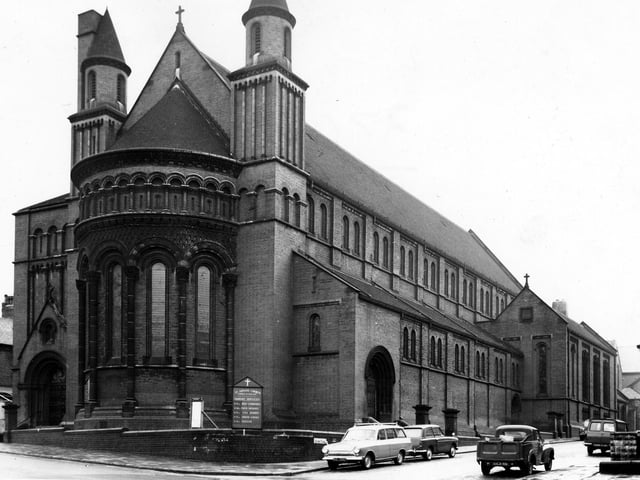 St Aidan's Church on Roundhay Road in April 1965 with Elford Place West on the right. Built in 1891-4 by R.J. Johnson & A.C. Hicks, St Aidan's joins Basilican form with Romanesque style. The image shows a double-apsed aisled basilica with a basilican chapel attached on the south eastern side. The interior of the church is famous for the large display of mosaics in the apse and on the concelli walls by Frank Brangwyn. Red brick and mosaic were chosen as construction materials as they were easy to maintain in Leeds polluted atmosphere. A large board in front of the church lists mass times.