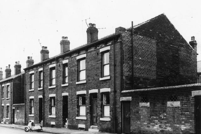 Three back-to-back terraced houses flanked by yards originally built to house the shared outside toilet. On the far left a child runs up the street while another child stands in the doorway of number 15. A moped is parked in the street outside number 15. This area was scheduled for demolition and the families relocated under a Leeds City Council slum clearance programme. Pictured in