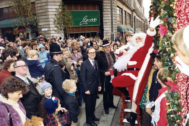 Crowds of people line The Headrow near Dortmund Square and the corner of Lewis's department store. The decorated float depicting a western steam engine can be glimpsed right. It was Lewis's entry in the 7th Lord Mayor's Parade and the overall winner. Santa waves to the crowds and behind him is a young lady dressed very regally later seen riding on the float. The gentleman seen centrally with spectacles is the General Manager of Lewis's, Steven Arundel. It was a blustery, showery day and many onlookers are wearing warm coats and hats. The view looks east towards Eastgate.