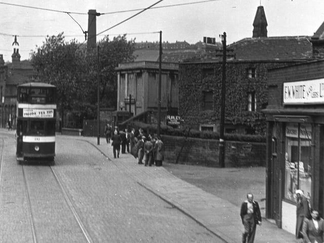 A tram travels along Meanwood Road  in JUn e 1955 with F W White, Drysalters, visible on the right. Next to this, covered in ivy and at an angle to the road, is Bog Hall. This house was adjacent to the ancient sulphur spa well and was possibly named for the bog land which typically produced sulphur springs. The spire of St Michael's Church on Buslingthorpe Lane can be seen above the roof. The light coloured building after Bog Hall is the transformer building for the trams on Meanwood Road, while the chimney to the left of this is in the Carr Mills complex on Buslingthorpe Lane. These buildings have all since been demolished leaving the white building on the far left as the only building in the image still standing. This is the Primrose pub at the junction of Meanwood Road and Buslingthorpe Lane.
